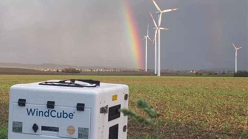 A WindCube ground LiDAR standing in a field after rain, wind turbines and a rainbow behind it