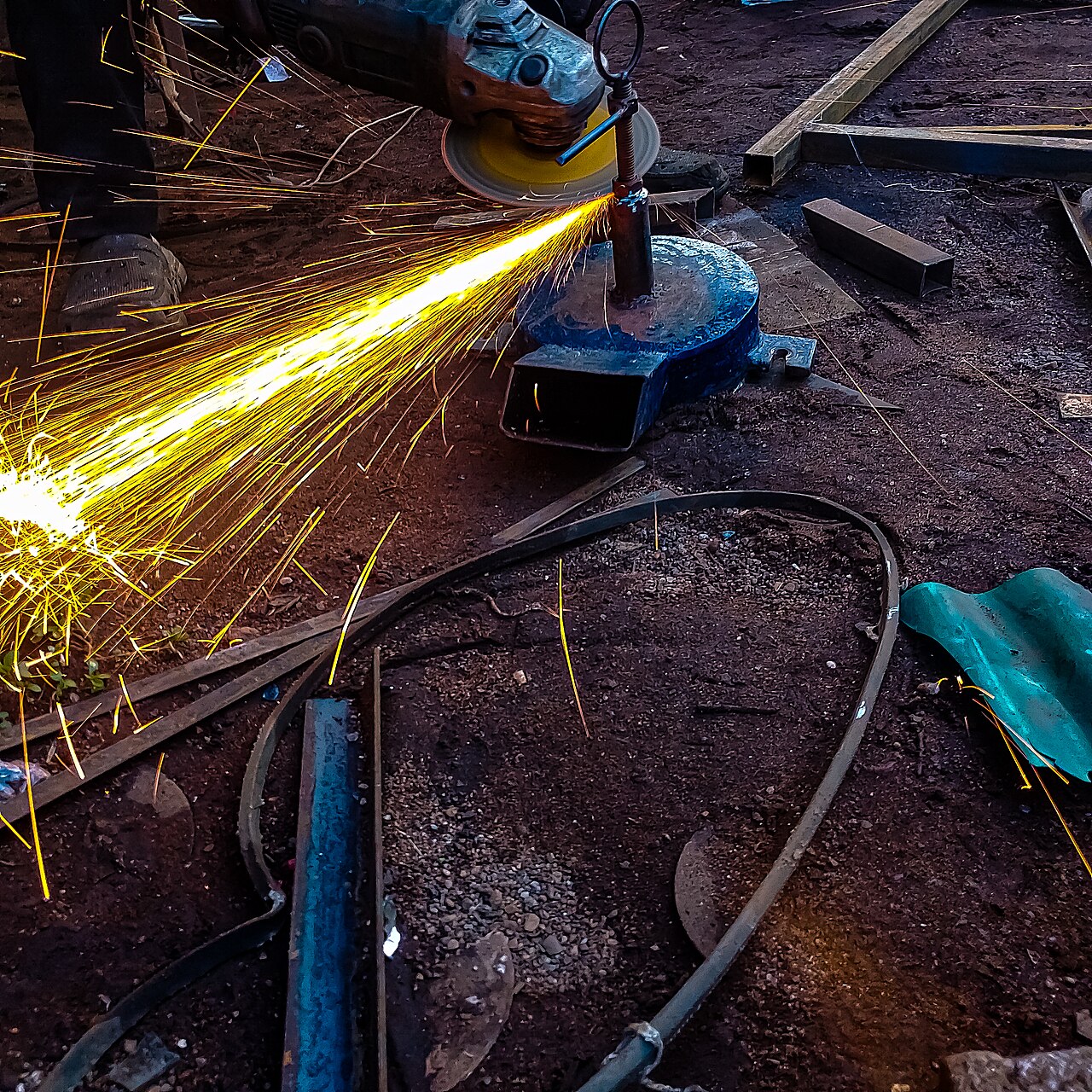 Steel being cut in the fabrication workshop — an angle grinder throwing a spray of sparks off a mast box-section, with steel stock laid out around the work