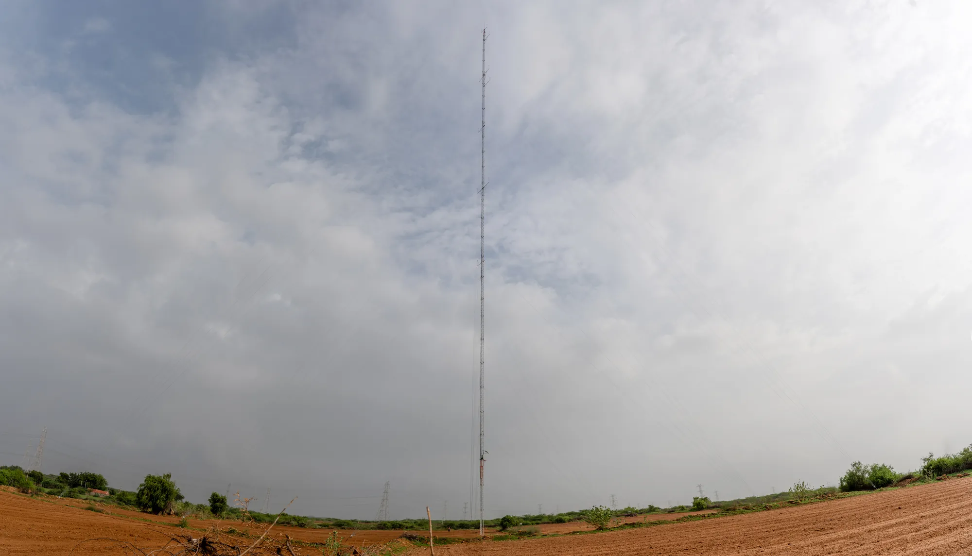 A tall guyed lattice met mast standing in an open field under a wide monsoon sky, power lines on the horizon